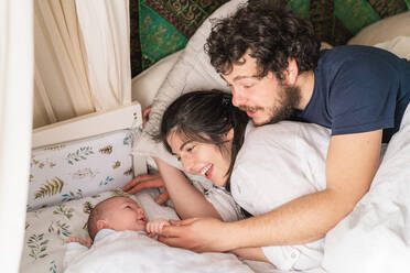 Von oben von fröhlichen Eltern, die mit einem süßen kleinen Kind interagieren, während es zu Hause im Bett liegt - ADSF24726