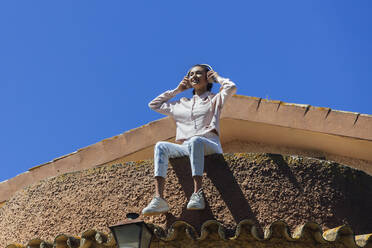 Smiling young woman listening music while sitting on roof - JRVF01006