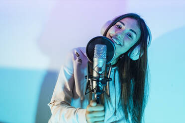 Smiling female singer wearing headphones singing in front of wall at studio - JRVF01030