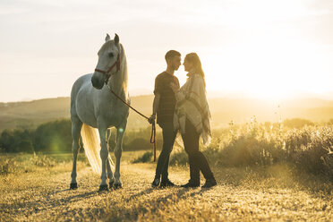 Romantisches junges Paar mit Pferd bei Sonnenuntergang auf einem Feld stehend - DAMF00804