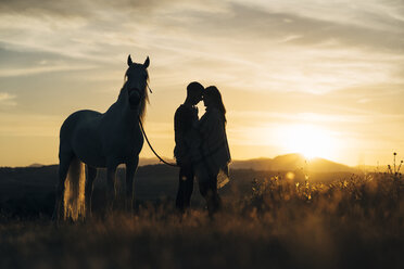 Silhouette eines Paares neben einem Pferd auf einem Feld bei Sonnenuntergang - DAMF00809