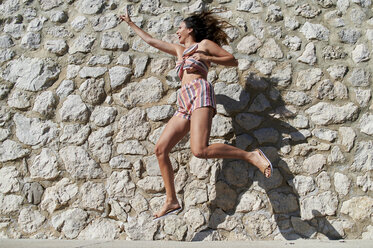 Woman gesturing while jumping by stone wall - KIJF03975