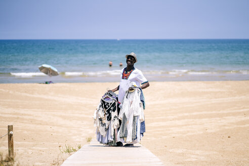 Männlicher Verkäufer steht auf der Strandpromenade und verkauft Kleidung am Strand an einem sonnigen Tag - OCMF02141