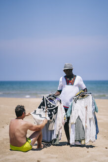 Mann kauft Kleidung von einem männlichen Verkäufer am Strand an einem sonnigen Tag - OCMF02142