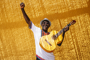 Fröhlicher Mann mit erhobener Hand und Gitarre vor einer gelben Wand an einem sonnigen Tag - OCMF02148
