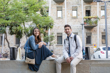 Smiling tourist couple sitting on concrete retaining wall - EIF01252