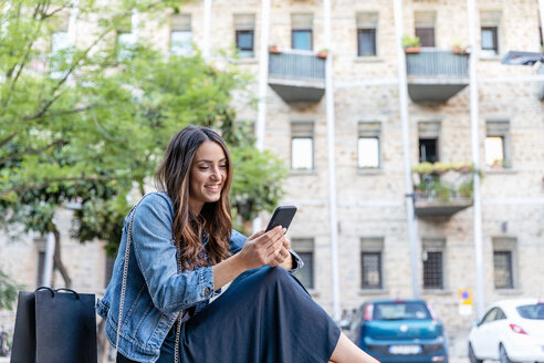 Lächelnde weibliche Touristin, die ihr Smartphone benutzt, während sie in der Stadt sitzt - EIF01256