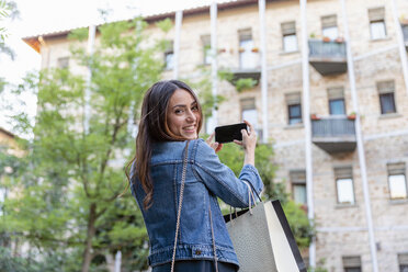 Lächelnder weiblicher Tourist mit Einkaufstaschen, der ein Selfie mit seinem Smartphone in der Stadt macht - EIF01261