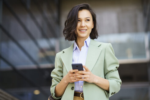 Lächelnde Geschäftsfrau, die wegschaut und ihr Smartphone vor einem Bürogebäude hält - JSMF02301