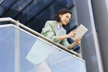 Lächelnde weibliche Fachkraft, die ein digitales Tablet benutzt, während sie sich am Geländer eines Bürogebäudes abstützt - JSMF02306