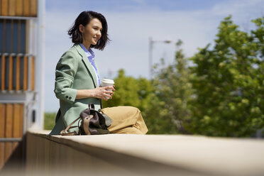 Geschäftsfrau sitzt auf einer Stützmauer und hält einen wiederverwendbaren Kaffeebecher an einem sonnigen Tag - JSMF02321