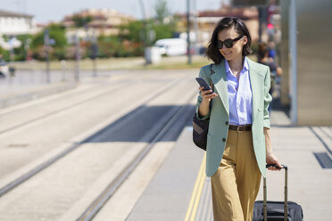 Berufstätige Frau, die ein Smartphone benutzt, während sie mit einem Koffer am Bahnhof spazieren geht - JSMF02326