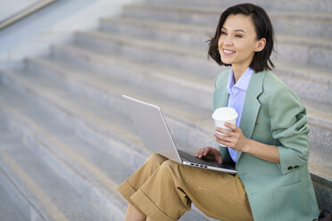 Smiling businesswoman with laptop looking away while sitting on steps - JSMF02329