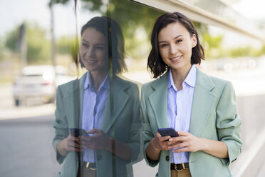Schöne Geschäftsfrau mit Smartphone an Glaswand gelehnt - JSMF02332
