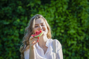 Lächelnde junge Frau mit Wassermelonenscheibe im Garten an einem sonnigen Tag - EIF01284