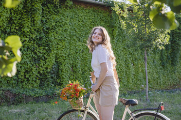 Cheerful young woman standing with bicycle in front of green ivy hedge - EIF01296