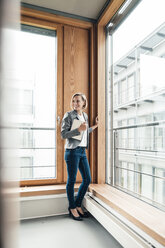 Businesswoman with digital tablet smiling while standing by office window - JOSEF04789