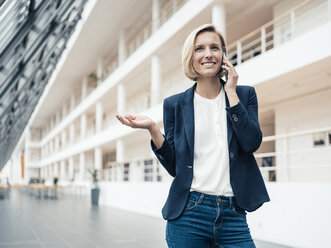 Businesswoman smiling while talking on mobile phone at office lobby - JOSEF04797