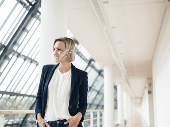 Businesswoman with transparent face mask standing at office corridor - JOSEF04802