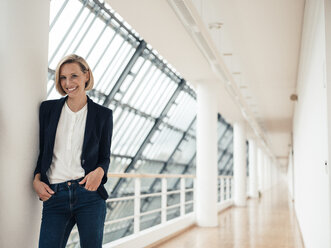 Female entrepreneur with hands in pockets standing at office corridor - JOSEF04803