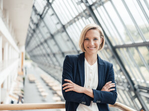 Smiling businesswoman with arms crossed leaning on railing in office - JOSEF04806