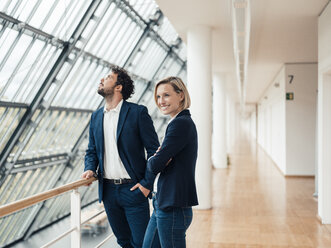 Smiling businesswoman with arms crossed standing by colleague at office corridor - JOSEF04834