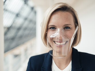 Smiling businesswoman with transparent protective face mask in office - JOSEF04849