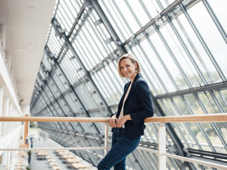 Female entrepreneur smiling while leaning on railing in office - JOSEF04851