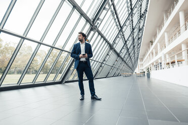 Male entrepreneur holding mobile phone while standing at office lobby - JOSEF04862