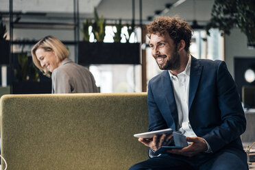 Lächelnder Geschäftsmann, der ein digitales Tablet hält, während er mit einem Kollegen im Hintergrund im Büro sitzt - JOSEF04907