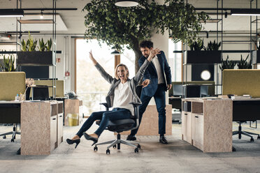 Cheerful businesswoman sitting on chair with colleague standing in background at office - JOSEF04908