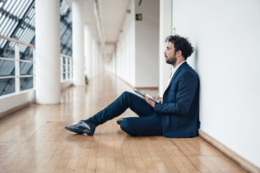 Businessman with digital tablet looking away while sitting at office corridor - JOSEF04920