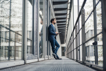 Thoughtful businessman with digital tablet standing at office corridor - JOSEF04925