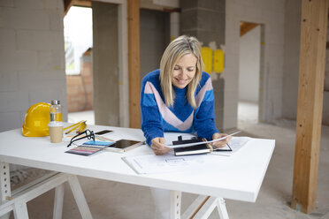 Lächelnde Architektin, die einen Bauplan prüft, während sie sich in einer Wohnung auf einen Tisch stützt - HMEF01288