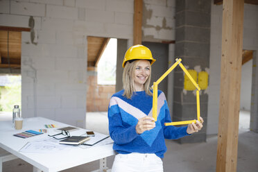 Lächelnde Bauunternehmerin mit Schutzhelm, die mit einem Zollstock in einer Wohnung ein Haus formt - HMEF01299