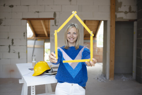Lächelnde weibliche Fachkraft, die durch einen Zollstock in Form eines Hauses auf einer Baustelle schaut - HMEF01302