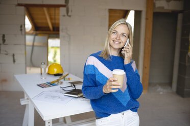Lächelnde Bauunternehmerin mit Einweg-Kaffeebecher, die mit ihrem Handy telefoniert und sich auf einen Tisch auf der Baustelle stützt - HMEF01307