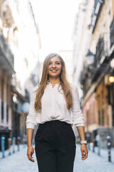 Smiling young woman with blond hair walking at alley - DAMF00824