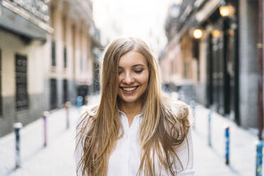 Fröhliche junge Frau mit blondem Haar in einer Gasse stehend - DAMF00828