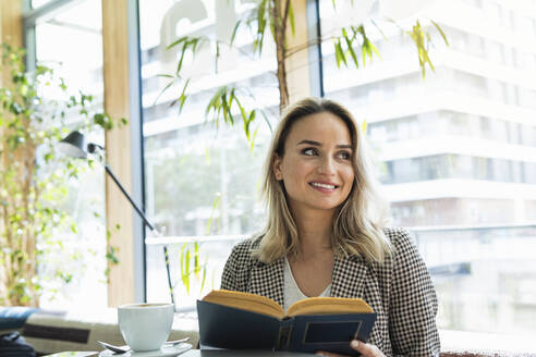 Lächelnde Geschäftsfrau mit Buch, die wegschaut, während sie im Café sitzt - PNAF01965