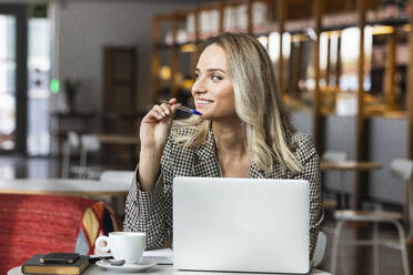 Lächelnde weibliche Fachkraft, die mit einem Laptop in einem Café sitzt und wegschaut - PNAF01967