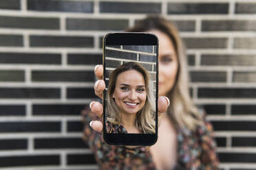 Junge Frau zeigt ein Foto auf ihrem Smartphone, während sie vor einer Wand steht - PNAF01979