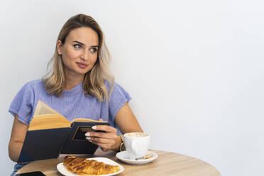 Junge Frau mit Buch, die wegschaut, während sie an einem Tisch vor einer Wand in einem Café sitzt - PNAF01989