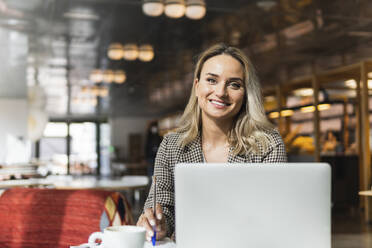 Lächelnde Geschäftsfrau hält einen Stift vor einem Laptop in einem Café - PNAF01998