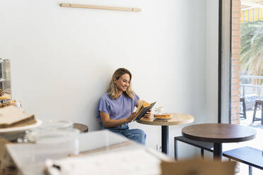 Smiling young woman reading book while sitting at table in coffee shop - PNAF02023