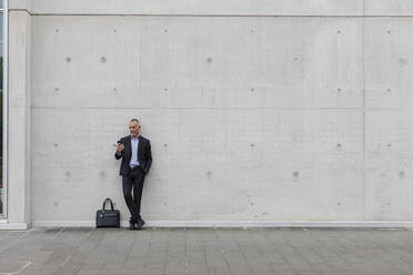 Männlicher Berufstätiger, der ein Smartphone benutzt, während er an einer Wand steht - EIF01303