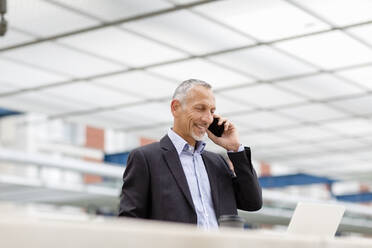 Lächelnder Geschäftsmann, der mit seinem Smartphone telefoniert und auf einen Laptop am Bahnhof schaut - EIF01326