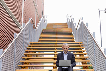 Männlicher Berufstätiger arbeitet an einem Laptop, während er auf einer Treppe in der Nähe eines Gebäudes sitzt - EIF01333
