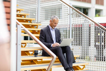 Männlicher Berufstätiger in Geschäftskleidung, der auf einer Treppe sitzend am Laptop arbeitet - EIF01335