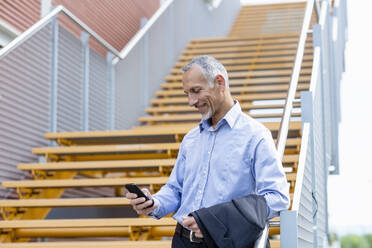 Geschäftsmann, der sein Smartphone benutzt, während er sich auf das Geländer in der Nähe der Treppe stützt - EIF01338